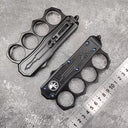 Two closed black knuckle duster OTF knives, one showing the front and one showing the back, displayed next to a metal ruler for size reference on a grey textured surface.
