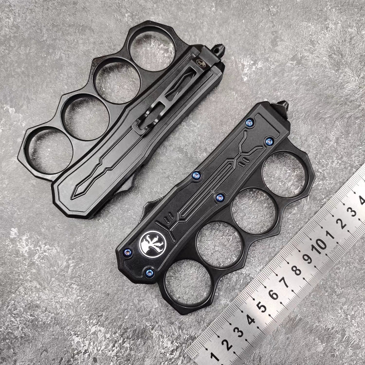 Two closed black knuckle duster OTF knives, one showing the front and one showing the back, displayed next to a metal ruler for size reference on a grey textured surface.