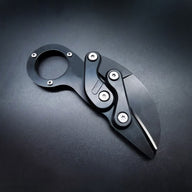 An open black mechanical folding karambit knife with a finger ring handle and articulated arms, displayed on a dark background.