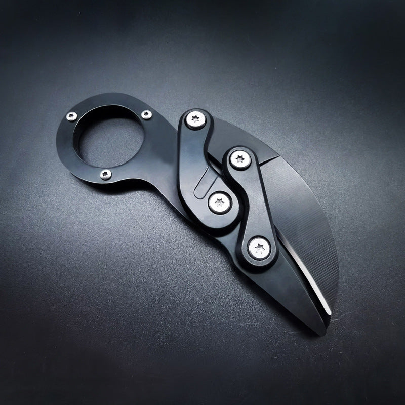 An open black mechanical folding karambit knife with a finger ring handle and articulated arms, displayed on a dark background.