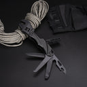 A black folding adjustable wrench multi-tool with multiple blades and tools deployed, displayed on a dark surface next to a coil of paracord and a black nylon sheath.
