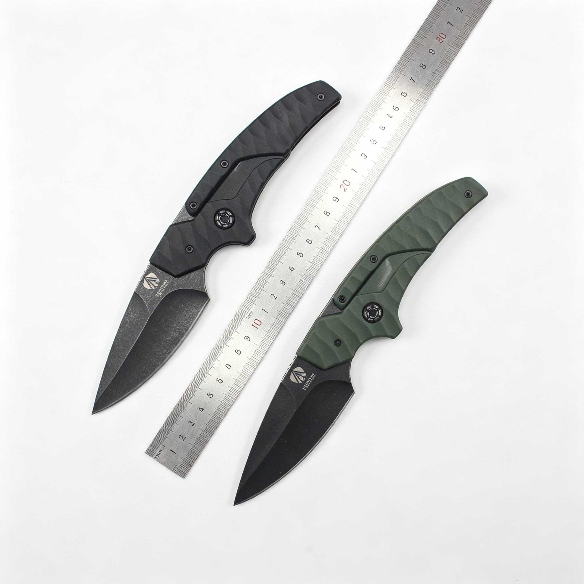 Two open folding knives with black blades and textured handles, one black and one olive green, displayed with a metal ruler between them on a white background for size reference.