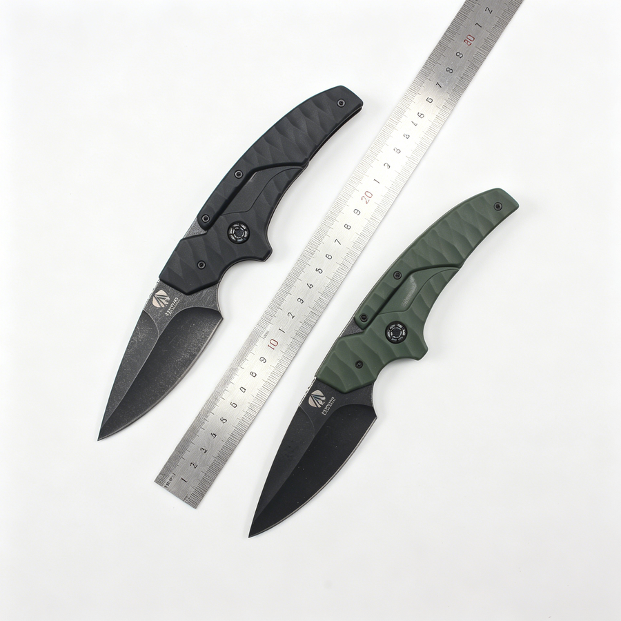 Two open folding knives with black blades and textured handles, one black and one olive green, displayed with a metal ruler between them on a white background for size reference.