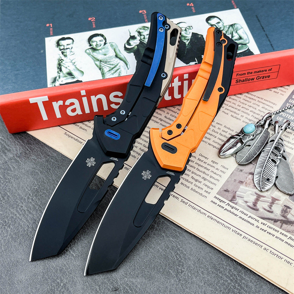 Two open tactical folding knives with black blades. The left knife features a black and tan handle with a blue pocket clip and button. The right knife features an orange handle with black accents. They are displayed on a red "Trainspotting" book next to silver feather pendants on a grey textured surface.
