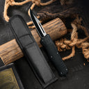A black OTF knife with a textured handle and silver blade, displayed next to a black nylon sheath against a rustic background of wood and rope.