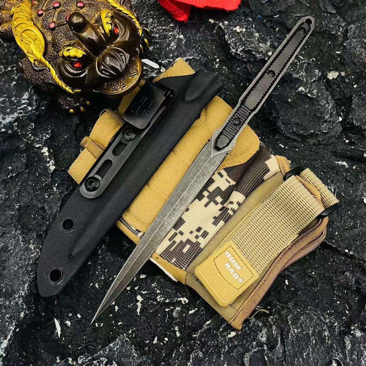 An open slim folding knife with a black blade displayed with its black sheath, a camo pouch, and a gold decorative statue on a rough rocky surface.