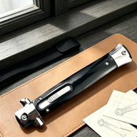 A closed stiletto knife with a black handle and silver hardware, resting on a brown leather notebook next to a black sheath straps and pencil sketches.