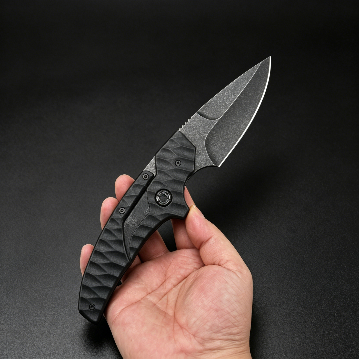 A hand holding an open black tactical folding knife with a textured handle and a dark stonewashed blade, displayed against a dark background.