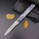  An open blue digital camo folding knife next to its black nylon pouch and dry autumn leaves on a textured black surface.