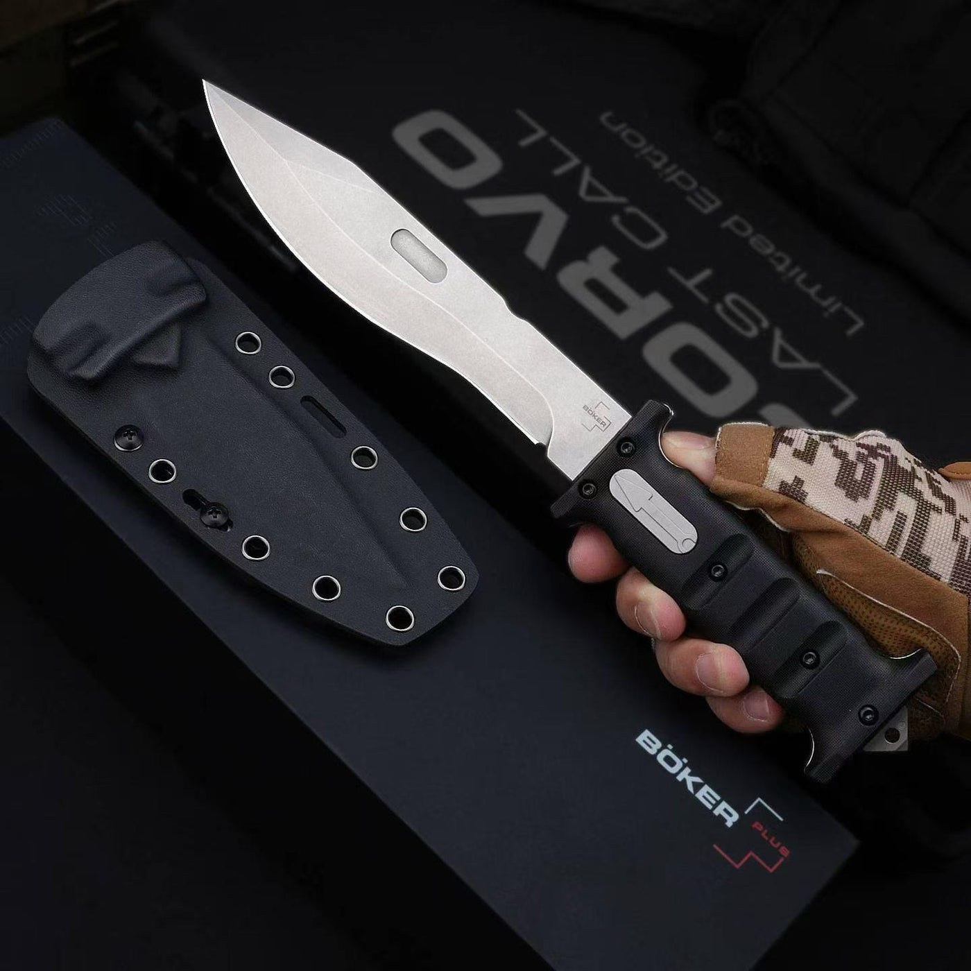 A hand wearing a brown and tan camo tactical glove holding an open Böker Plus Last Corvid folding knife with a silver blade and black handle, displayed next to a black hard sheath and packaging box.