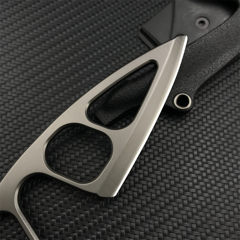 A close-up of the silver stainless steel tip of a Böker Plus skeletonized neck knife, resting on a black leather sheath against a dark carbon fiber textured background.