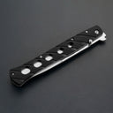 A closed black folding knife featuring a handle with circular cutouts, a silver thumb stud, and a silver blade edge visible along the bottom, displayed on a dark gray background.