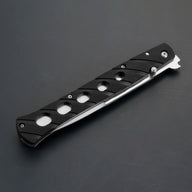 A closed black folding knife featuring a handle with circular cutouts, a silver thumb stud, and a silver blade edge visible along the bottom, displayed on a dark gray background.