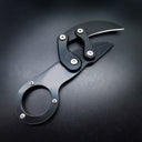 A closed black mechanical folding karambit knife with a finger ring handle, articulated arms, and visible star screws, displayed on a dark textured surface.