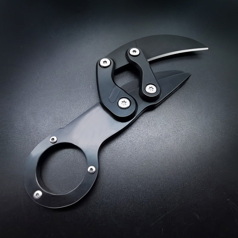 A closed black mechanical folding karambit knife with a finger ring handle, articulated arms, and visible star screws, displayed on a dark textured surface.