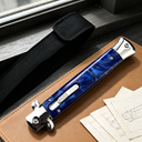 A closed stiletto knife with a blue resin handle and silver hardware, resting on a leather notebook next to a black nylon sheath and technical sketches.