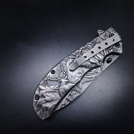 A closed folding knife featuring deep relief engravings of a deer, mountains, and forest trees on the handle and blade, equipped with a silver pocket clip, displayed on a dark background.