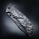 A closed folding knife with deep relief engraving of several deer and forest scenery on the handle and blade, displayed on a dark textured background.