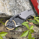 A closed folding knife with a grey handle and gold gear pivot, resting on a stone with green leaves and a red box outdoors.