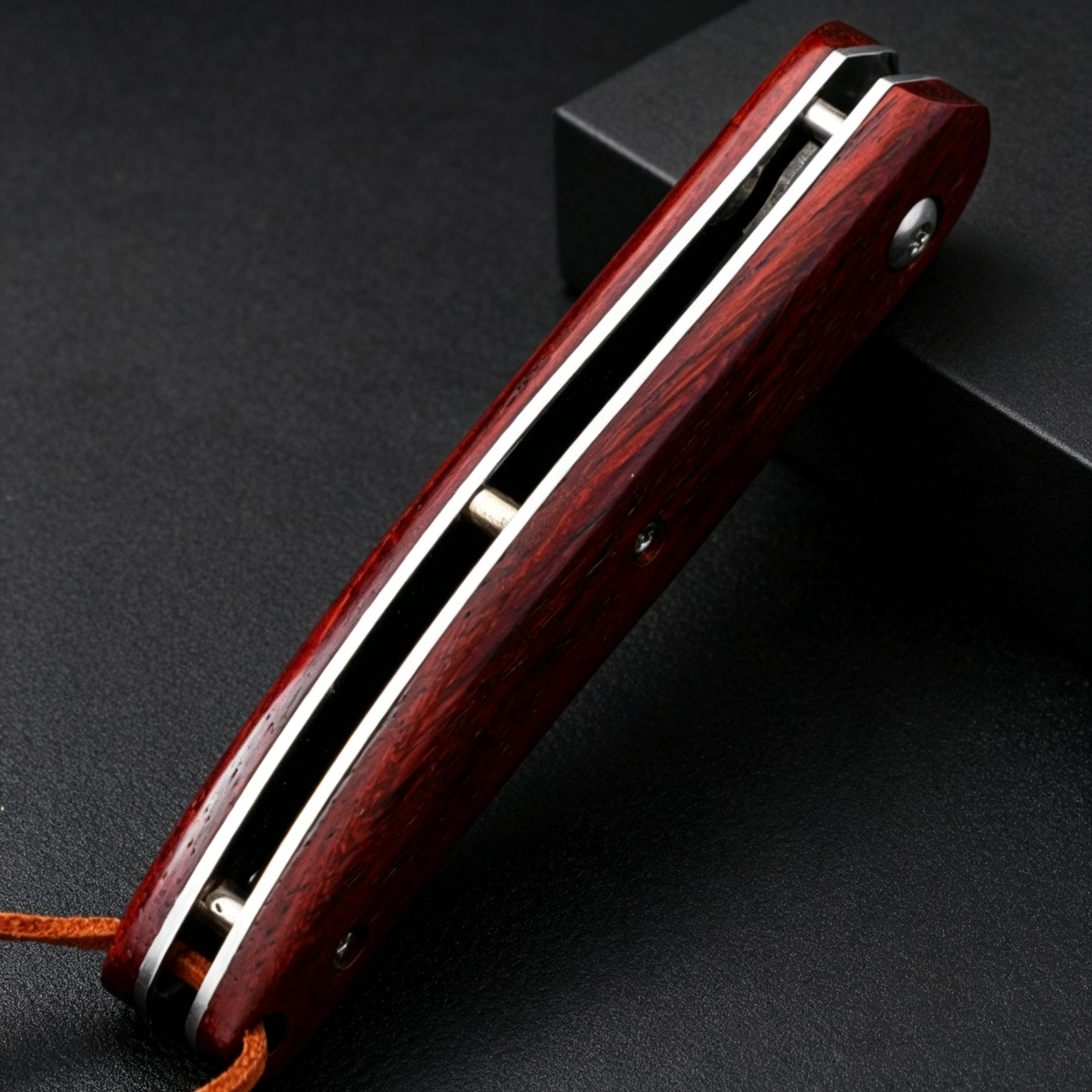 A closed folding knife with a polished red wooden handle and silver liners, featuring a brown lanyard at the end, resting against a black box on a dark textured background.
