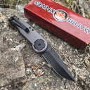 A closed folding knife with a grey handle and black pocket clip, resting on a red packaging box outdoors.