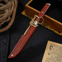 A closed M390 folding knife displayed alongside its wooden sheath equipped with a brass locking mechanism and key ring, surrounded by rope and a wooden display box.