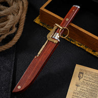 A closed M390 folding knife displayed alongside its wooden sheath equipped with a brass locking mechanism and key ring, surrounded by rope and a wooden display box.