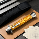 A closed stiletto knife featuring a bright orange resin handle and silver hardware, resting on a leather notebook next to black sheath straps and technical drawings.