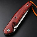 A closed folding knife featuring a red wooden handle, silver screws, a visible damascus steel blade with patterns, and a brown leather lanyard, resting on a dark textured surface.