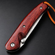 A closed folding knife featuring a red wooden handle, silver screws, a visible damascus steel blade with patterns, and a brown leather lanyard, resting on a dark textured surface.