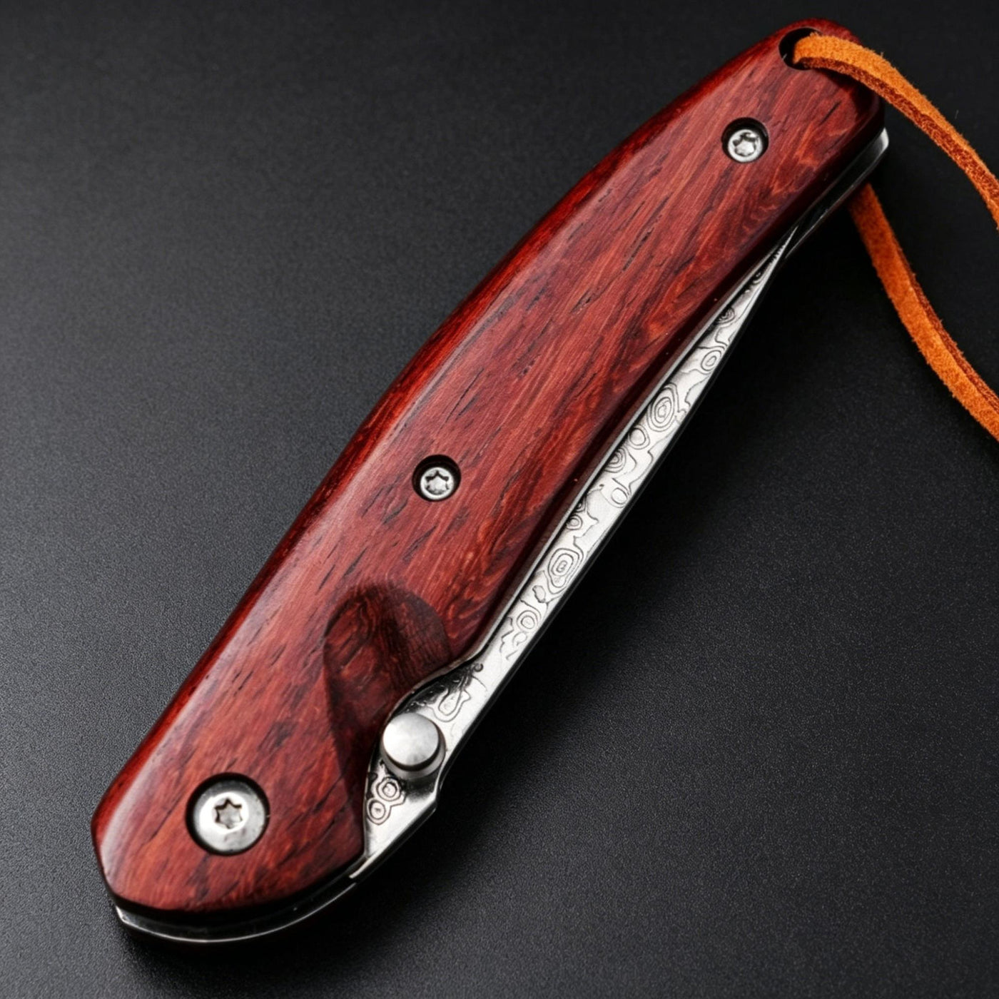 A closed folding knife featuring a red wooden handle, silver screws, a visible damascus steel blade with patterns, and a brown leather lanyard, resting on a dark textured surface.
