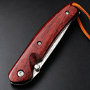 A closed folding knife featuring a red wooden handle, silver screws, a visible damascus steel blade with patterns, and a brown leather lanyard, resting on a dark textured surface.