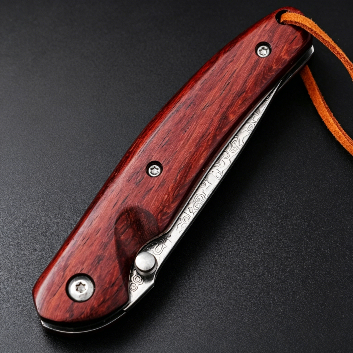 A closed folding knife featuring a red wooden handle, silver screws, a visible damascus steel blade with patterns, and a brown leather lanyard, resting on a dark textured surface.