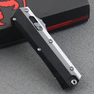 A close-up of a closed out-the-front knife with a silver and black handle, showing the firing button and screws, resting on a black box.