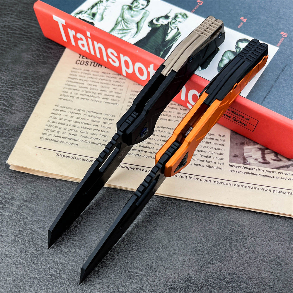 Two closed slim folding knives with black blades and push-button mechanisms. The top knife features a black and tan handle, and the bottom knife features a black and orange handle, resting on an open magazine page on a textured grey surface.