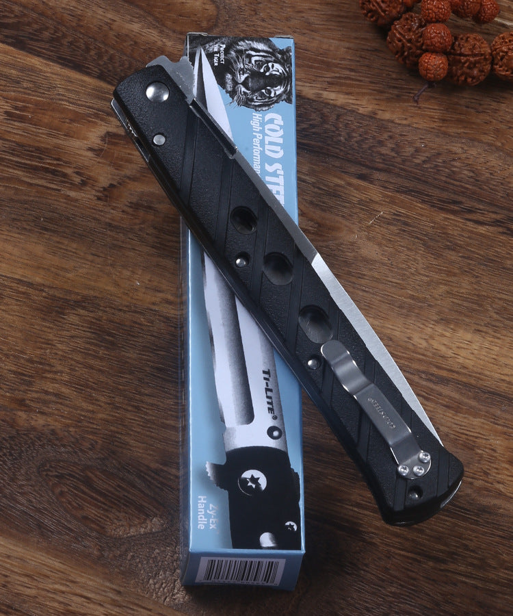 An open Cold Steel Ti-Lite folding knife with a black handle and silver pocket clip, resting on top of its blue packaging box on a wooden desk surface.