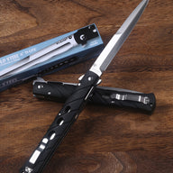 An open and a closed Cold Steel Ti-Lite folding knife with black handles, displayed next to a blue packaging box on a wooden desk.