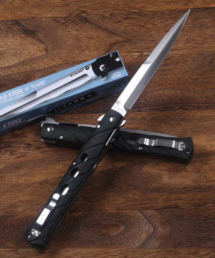 An open and a closed Cold Steel Ti-Lite folding knife with black handles, displayed next to a blue packaging box on a wooden desk.