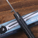 A partially open Cold Steel Ti-Lite folding knife with a black handle and silver blade, resting across its blue packaging box on a wooden table, showing the spine and pocket clip details.