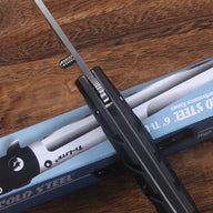 A partially open Cold Steel Ti-Lite folding knife with a black handle and silver blade, resting across its blue packaging box on a wooden table, showing the spine and pocket clip details.