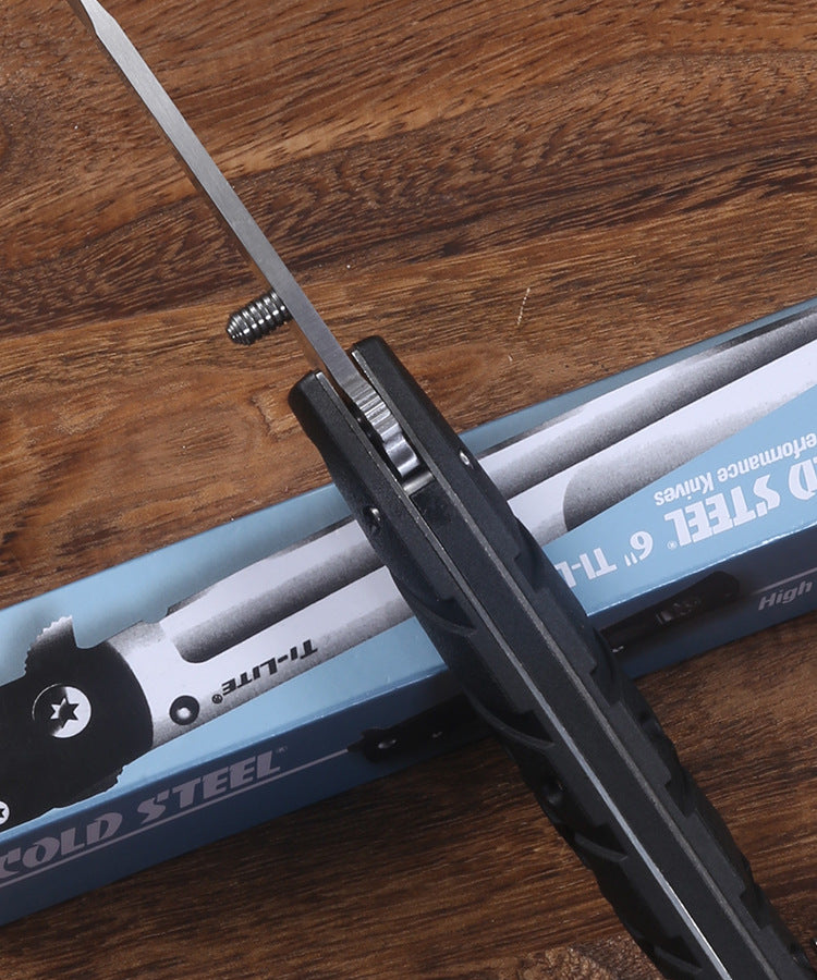 A partially open Cold Steel Ti-Lite folding knife with a black handle and silver blade, resting across its blue packaging box on a wooden table, showing the spine and pocket clip details.