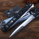 Two open Cold Steel Ti-Lite folding knives with black handles and silver blades, displayed on a blue packaging box next to a dark slate stone and brown beads on a wooden desk.