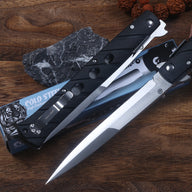 Two open Cold Steel Ti-Lite folding knives with black handles and silver blades, displayed on a blue packaging box next to a dark slate stone and brown beads on a wooden desk.