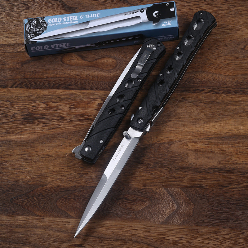An open and a closed Cold Steel Ti-Lite folding knife with black handles and silver blades, displayed next to their blue packaging box on a wooden desk.