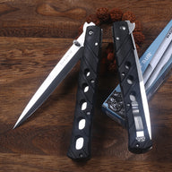 Two open Cold Steel Ti-Lite VI folding knives with black handles and silver blades, displayed on a wooden desk next to a decorative bead strand and the original packaging box.