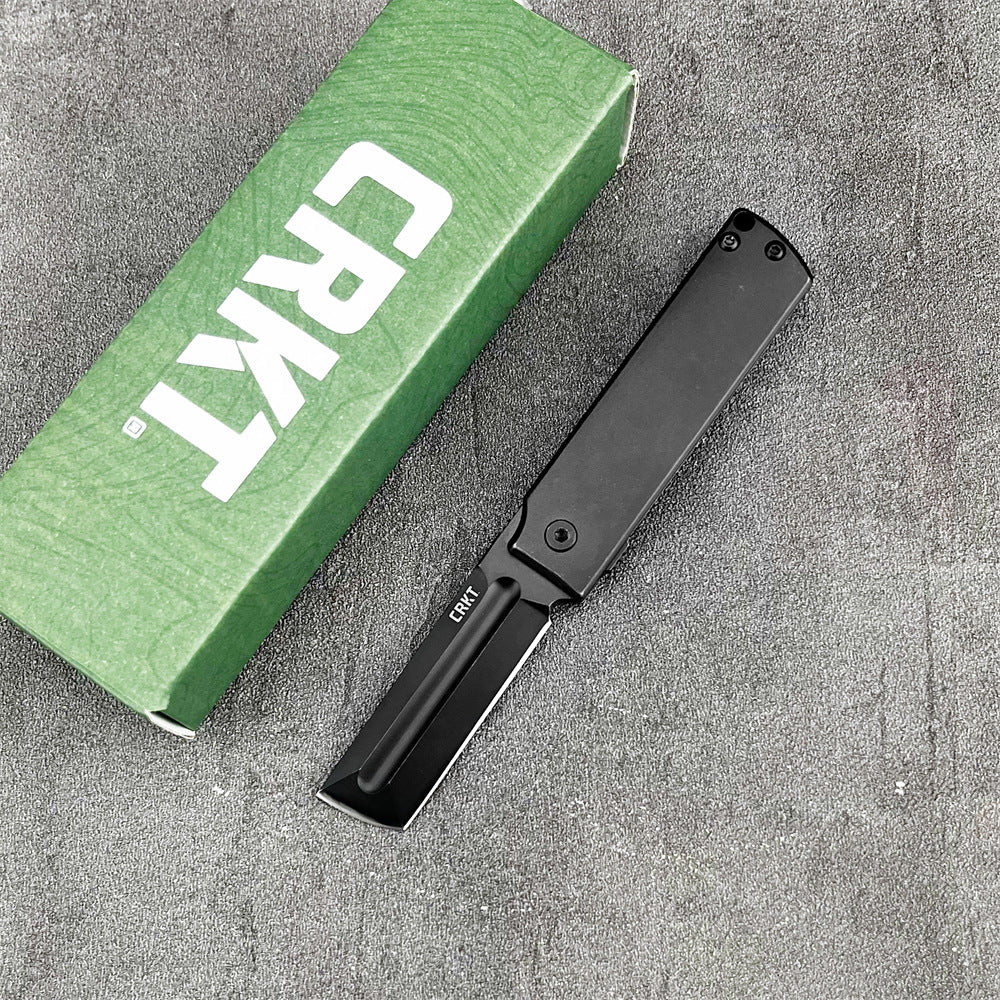 A closed CRKT folding knife with a black handle, displayed next to a green CRKT box on a grey textured surface.