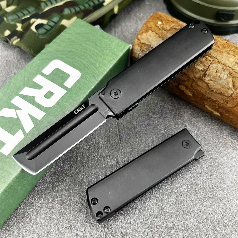 An open and a closed CRKT folding knife with black handles and tanto blades, displayed on a grey surface with a green box, wood log, and camo bag.