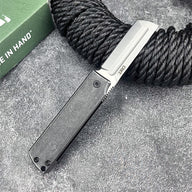 An open CRKT folding knife with a black handle and silver tanto blade, displayed on a rough grey surface next to a black rope and green CRKT box.