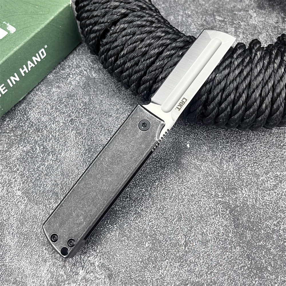 An open CRKT folding knife with a black handle and silver tanto blade, displayed on a rough grey surface next to a black rope and green CRKT box.