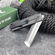 An open CRKT folding knife with a black handle and silver tanto blade, displayed on a grey surface with black rope and a green box.